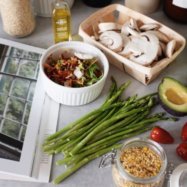 A selection of fresh vegetables and a cookbook in a kitchen setting, promoting healthy cooking.