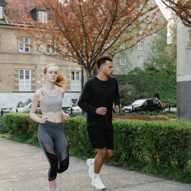 Two adults jogging on a sidewalk in an urban setting surrounded by spring foliage.