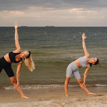 Two women exercising at a beach, performing yoga poses with the ocean in the background during sunset.
