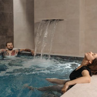 A couple relaxing in an indoor spa pool under a soothing waterfall effect.